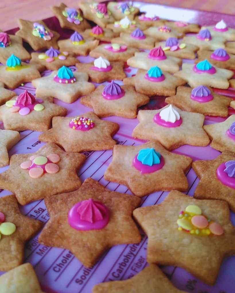Biscuits en forme d’étoile décorés avec glaçage coloré et bonbons, sur tapis de pâtisserie rose.