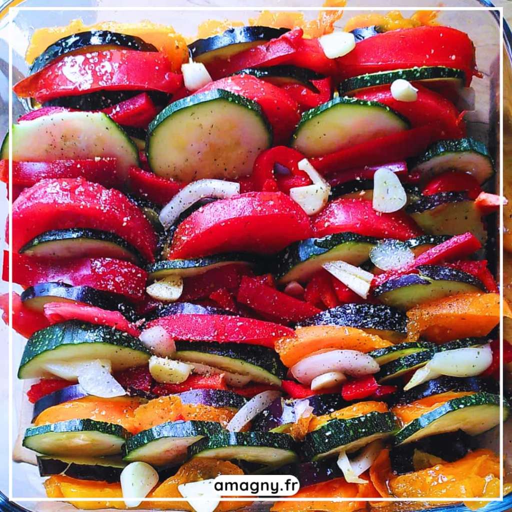 Plat de légumes tranchés — courgettes, tomates, poivrons et ail — disposés en tian coloré.