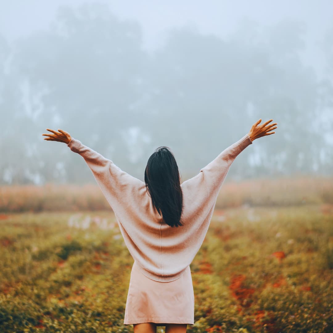 Femme de dos, bras ouverts, dans un paysage brumeux.