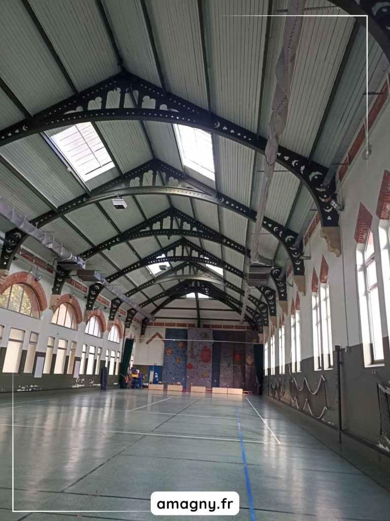 Gymnase spacieux avec charpente mĂ©tallique, grandes fenĂȘtres et mur dâescalade colorĂ©.
