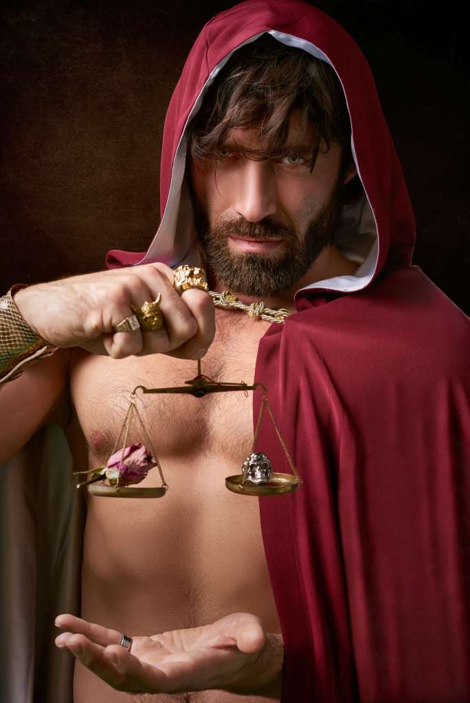 Homme portant une cape rouge tenant une balance avec une rose séchée et un crùne métallique.