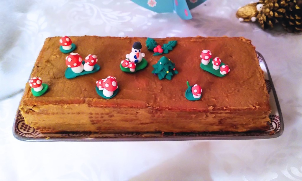 Gâteau rectangulaire décoré de figurines de Noël sur glaçage chocolat, posé sur plateau festif. 