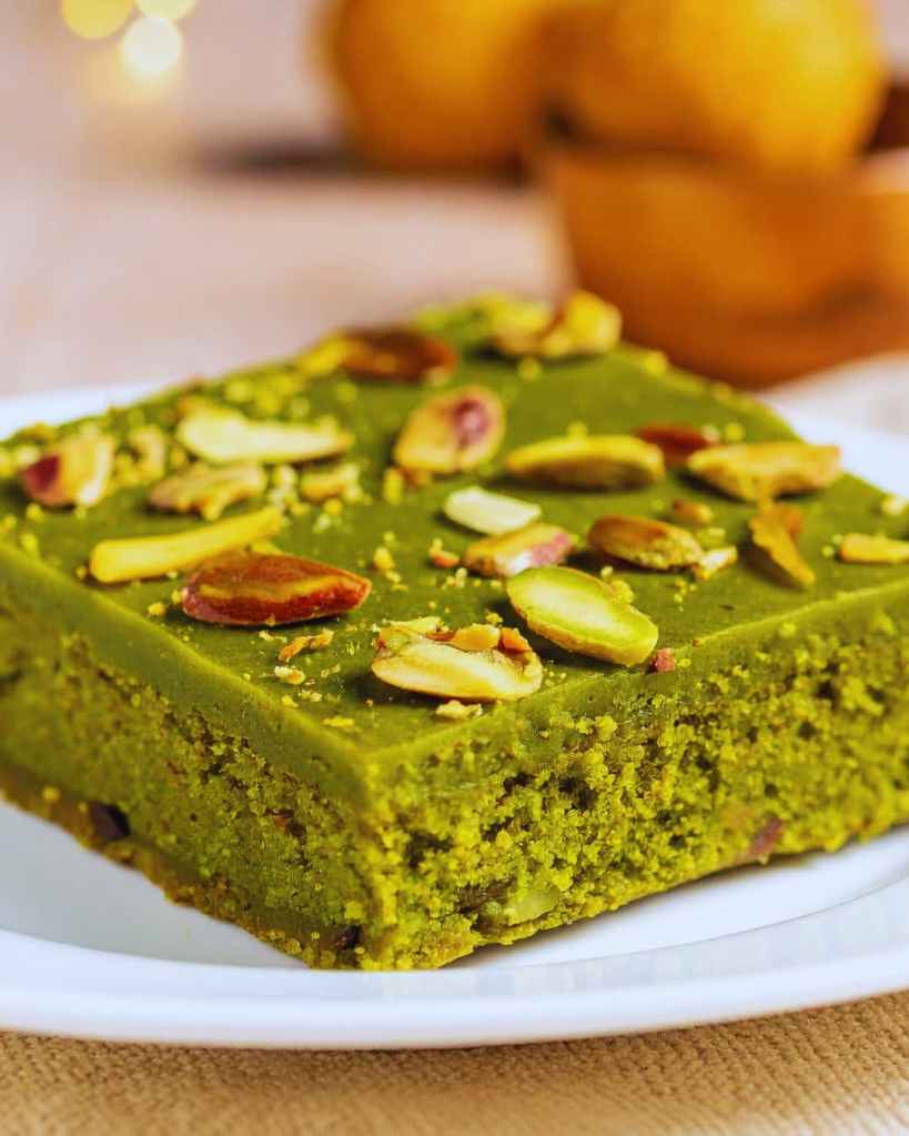 Tranche de cake vert au matcha, décorée de pistaches et d’amandes sur assiette blanche.