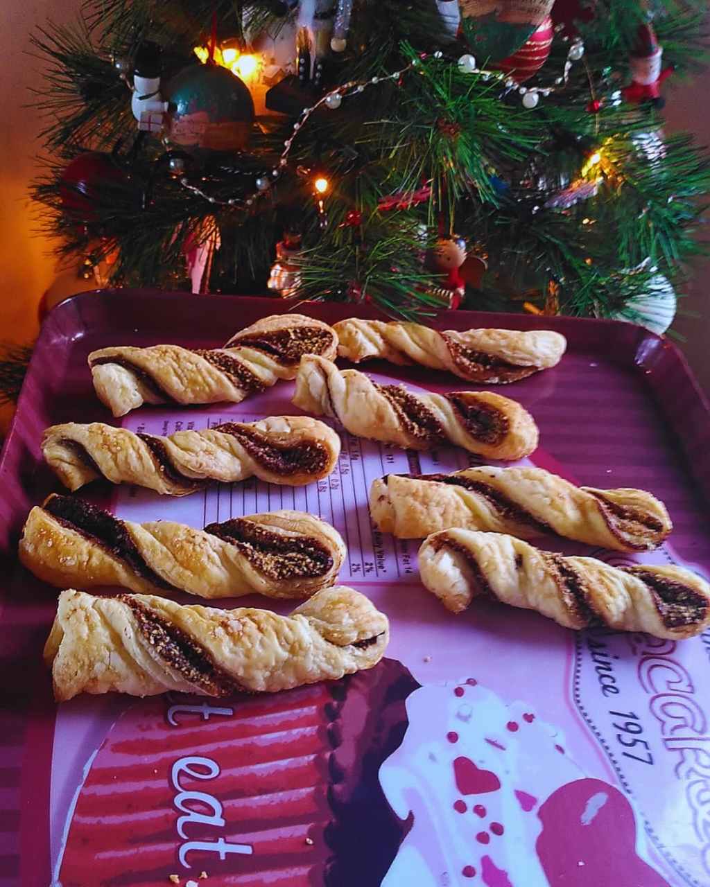 Torsades feuilletées dorées, présentées sur un plateau rouge devant un sapin décoré.