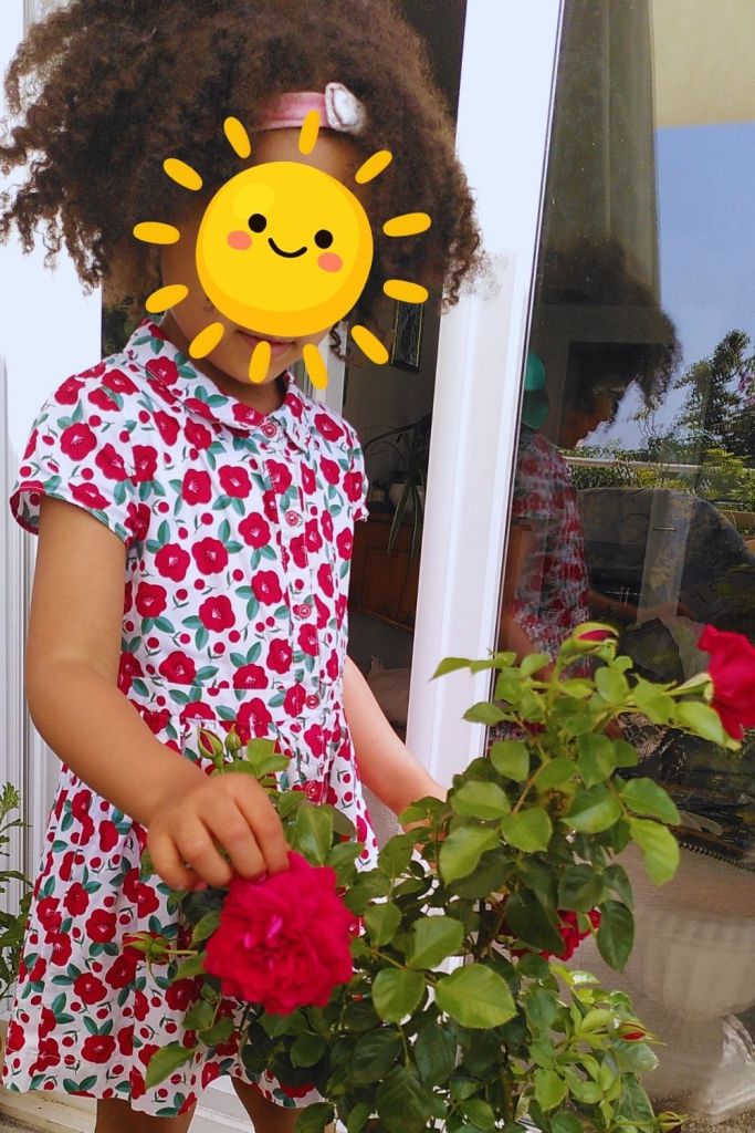 Enfant tenant une fleur de camélia rouge devant une porte vitrée, entouré de plantes.